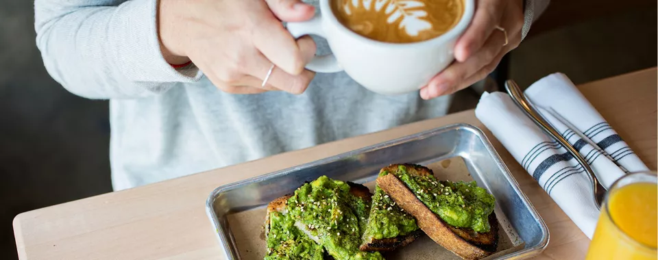 Person holding a cup of cappuccino with a plate of avocado toast on the table